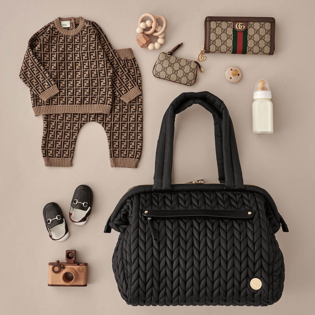 Paige Carryall Black from the front, showing front mommy pocket, herringbone quilting, gold hardware, and padded carry handles; laid flat with matching beige and black mom and baby items.