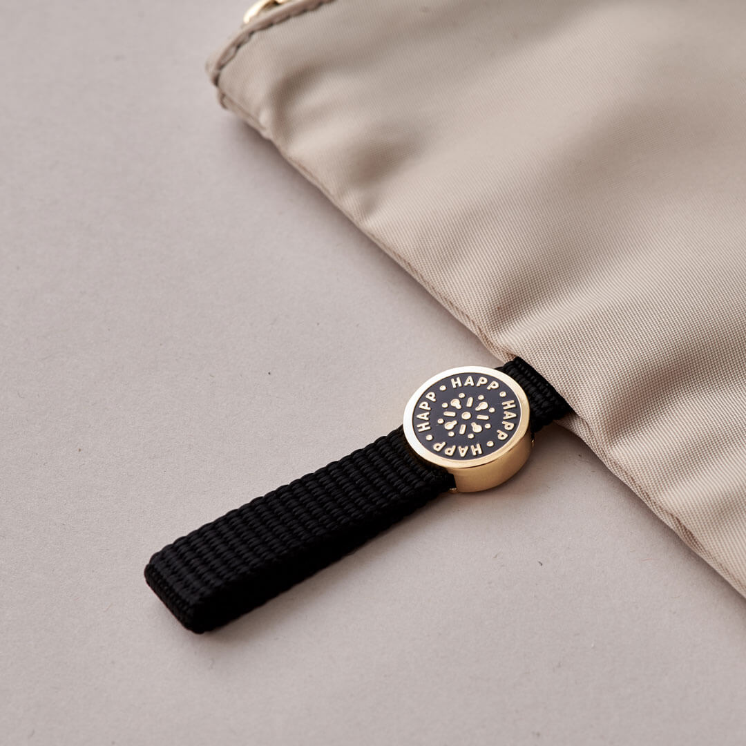 Closeup of the black pull tab and enamel logo while beige pouch is lying flat on a beige background.