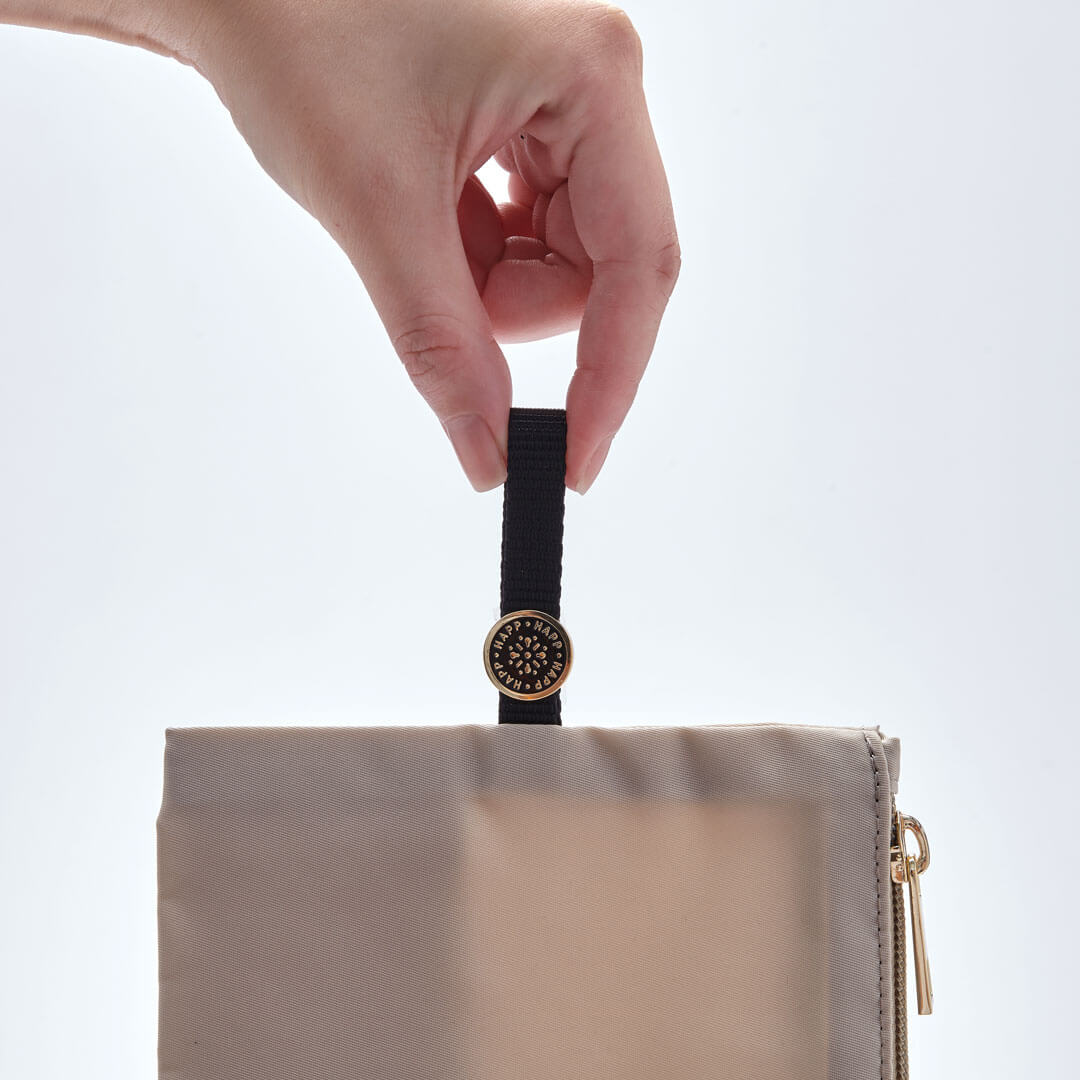 Closeup of a hand holding the black pull tab found on the Large and Medium Beige pouches, showing off the enamel HAPP logo also in black.