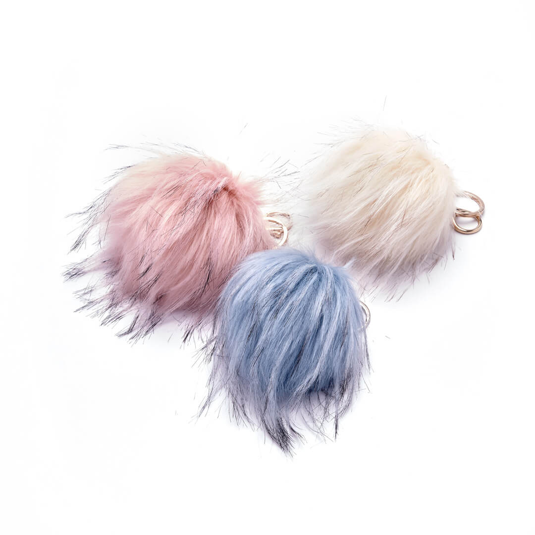 Three faux fur pouf bag charms in rose, blue, and ivory colors with keyring attachments.