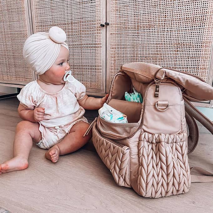 A baby girl in a pink top-knot bonnet with a pacifier sits beside a Levy Backpack Dusty Rose, which sits upright and open packed with baby items. The baby is looking at and touching the diaper bag.