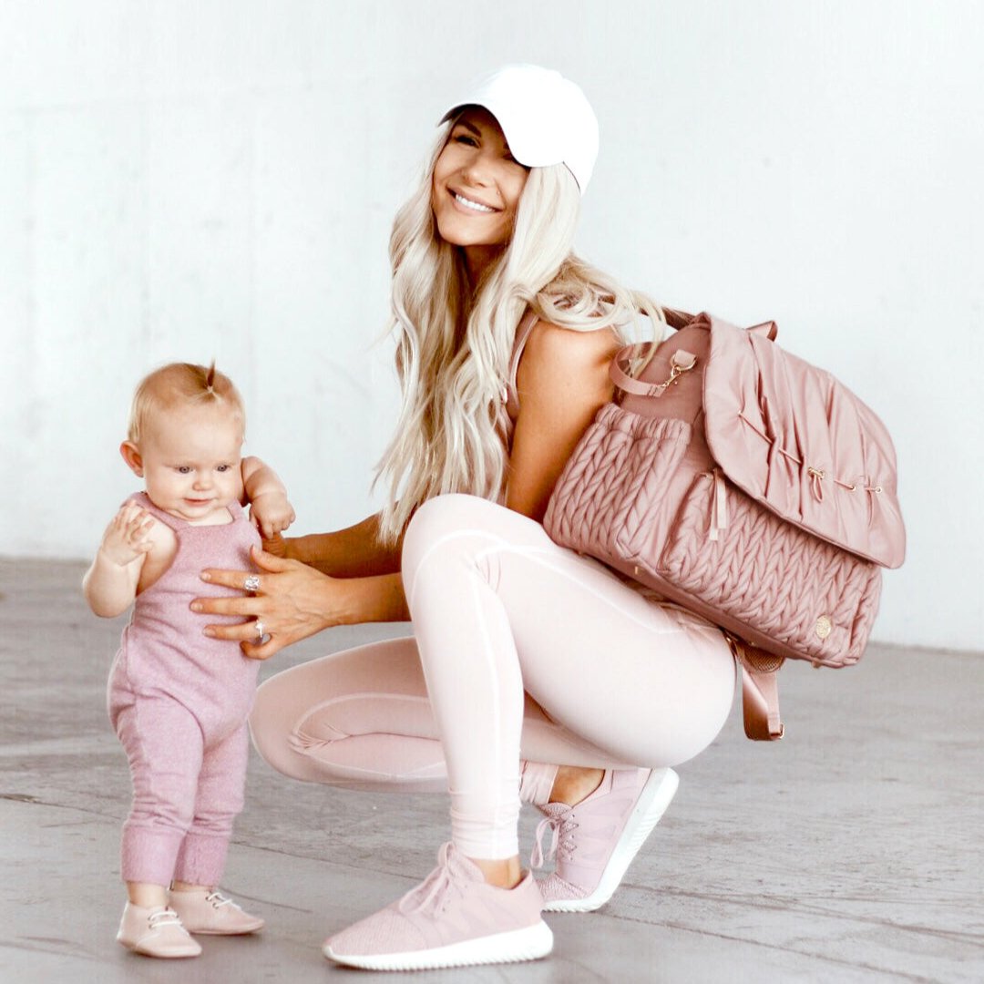 A stylish mom in pink athleisure crouching down to attend to her toddler, wearing the Levy Backpack Dusty Rose over one shoulder and smiling at the camera, showing the versatility of style of HAPP diaper bags.