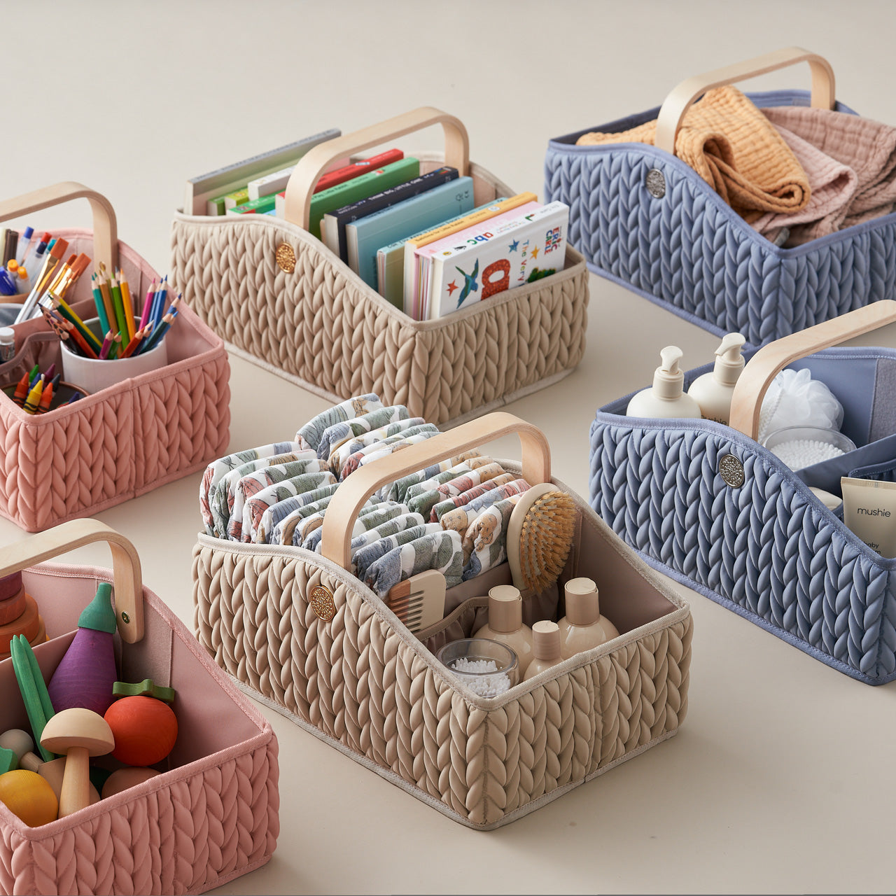 6 diaper caddies, 2 blue, 2 beige, 2 rose, standing upright in rows, packed with different themes: art supplies, toys, books, blankets, diapering items, and bathtime items.
