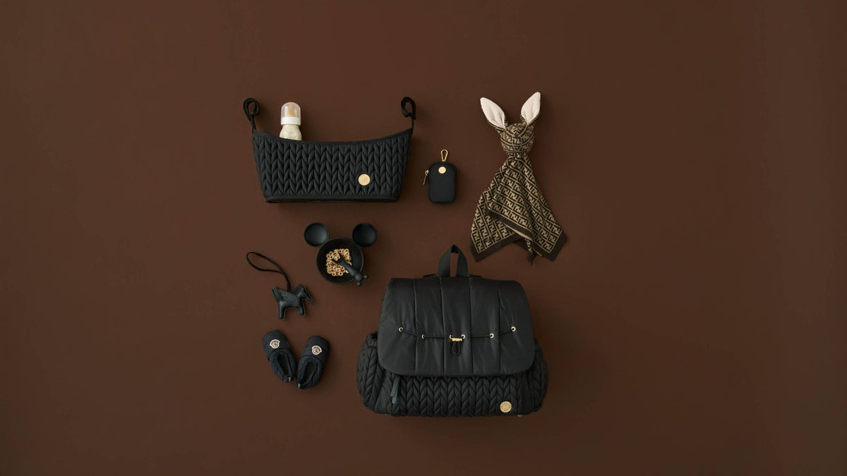 Flat lay image of HAPP Brand Levy Backpack, Stroller Organizer, and Clip-on Pouch in Classic Black, arranged with matching black baby shoes, keychain, bowl and spoon, and brown baby blanket on a warm brown background.