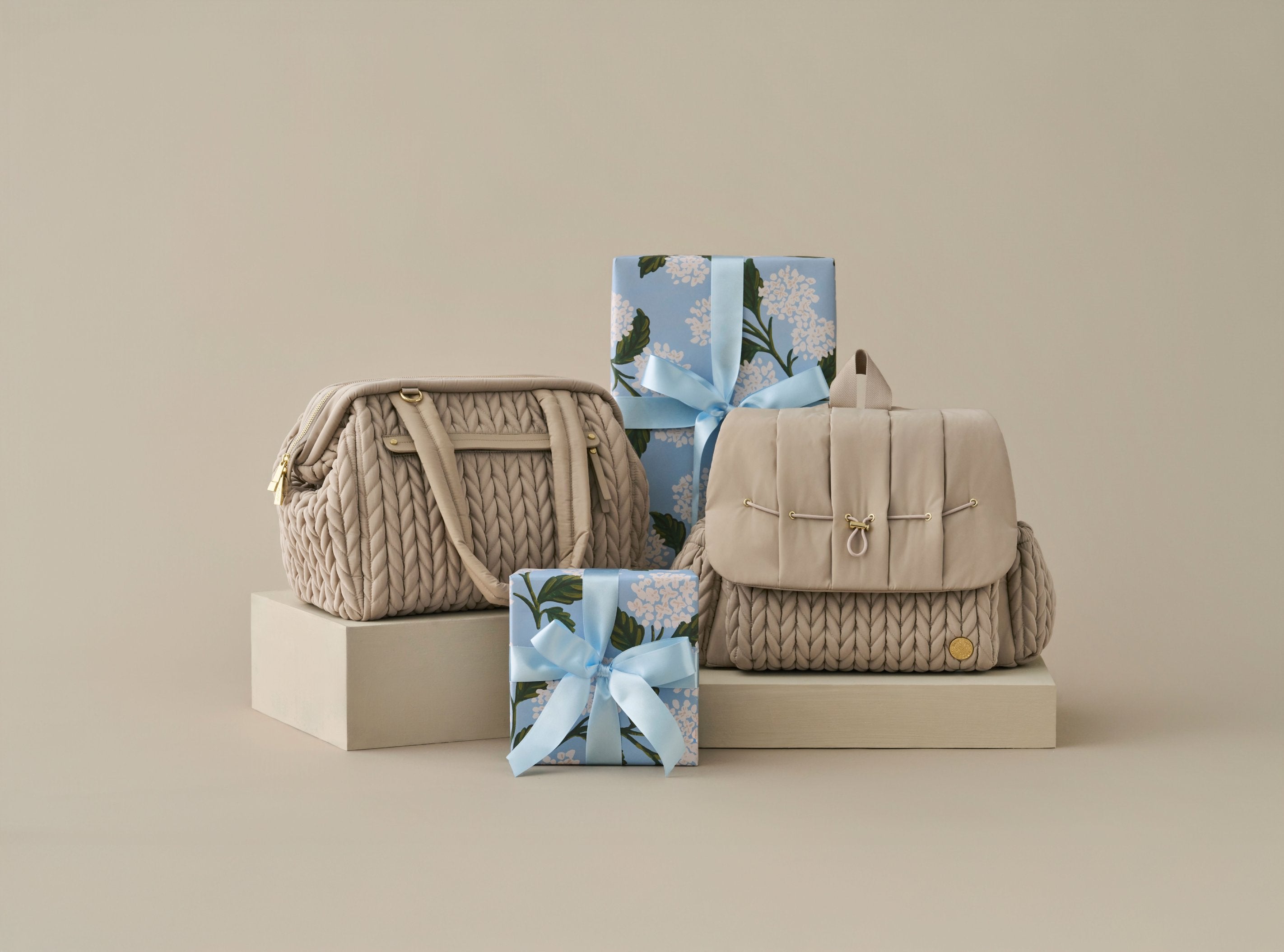 HAPP Brand Levy Backpack and Paige Carryall in Beige, standing upright beside gifts wrapped in blue paper, on a beige background.