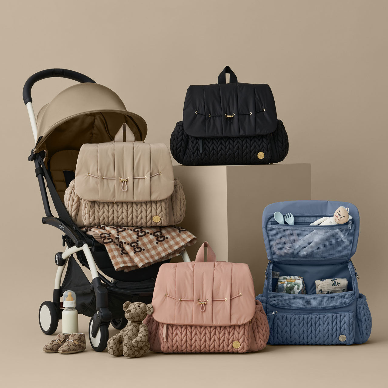 HAPP Brand Levy Backpack Diaper Bag shown in all four colors: Black, Beige, Rose, and Blue Herringbone, on a beige background. The Beige bag sits on a stroller and the Blue bag is shown open, displaying a well-organized interior. 