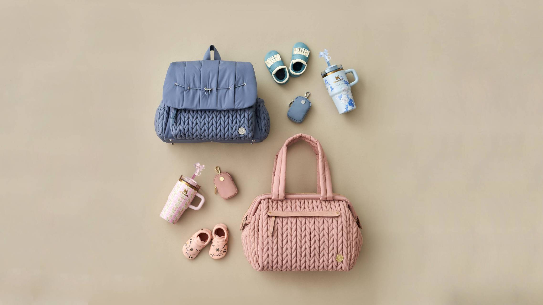HAPP Brand Levy Backpack Ash Blue and Paige Carryall Dusty Rose with matching blue and rose Clip-on Pouches, tumblers, and baby shoes on a beige background.