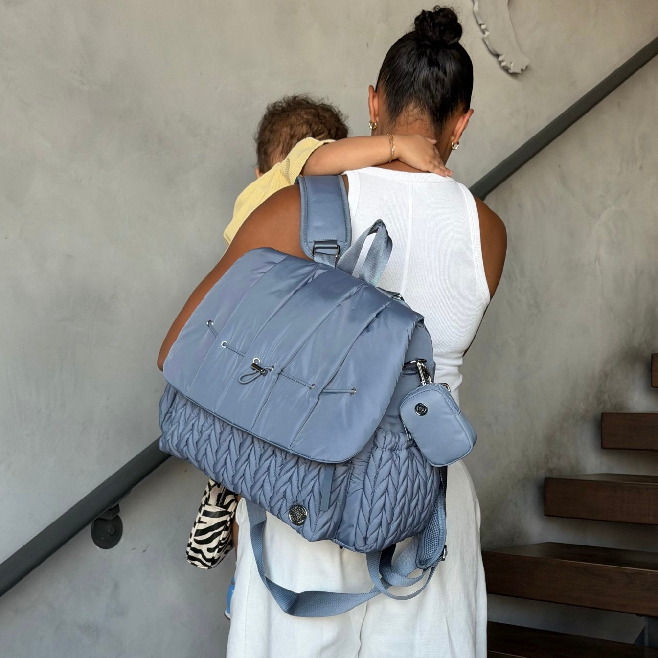 A mom carrying her baby and wearing the HAPP Brand Levy Backpack Diaper Bag in Ash Blue Herringbone, wearing a white dress and seen from behind walking up exterior stairs in front of a beige wall.