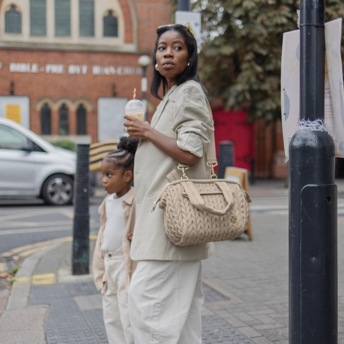 A stylish mom carrying a HAPP Brand Paige Mini Diaper Bag in Beige Herringbone, waiting to cross the street with her daughter. Both are wearing matching off-white outfits.